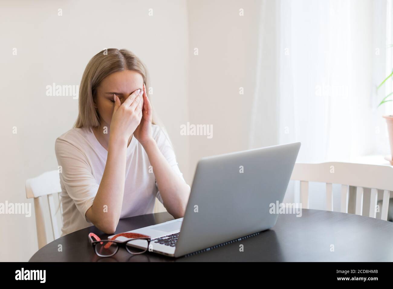 Fatiga, trabajo excesivo, cansancio. Mujer exhausta tiene un dolor de cabeza después de trabajar en el ordenador portátil, tocándola los ojos, tratando de concentrarse, sentada junto a la mesa Foto de stock