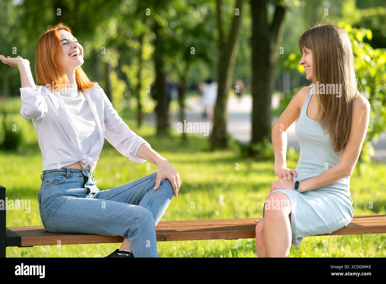 Dos jóvenes amigas sentadas en un banco en el parque de verano charlando felices y divirtiéndose. Foto de stock