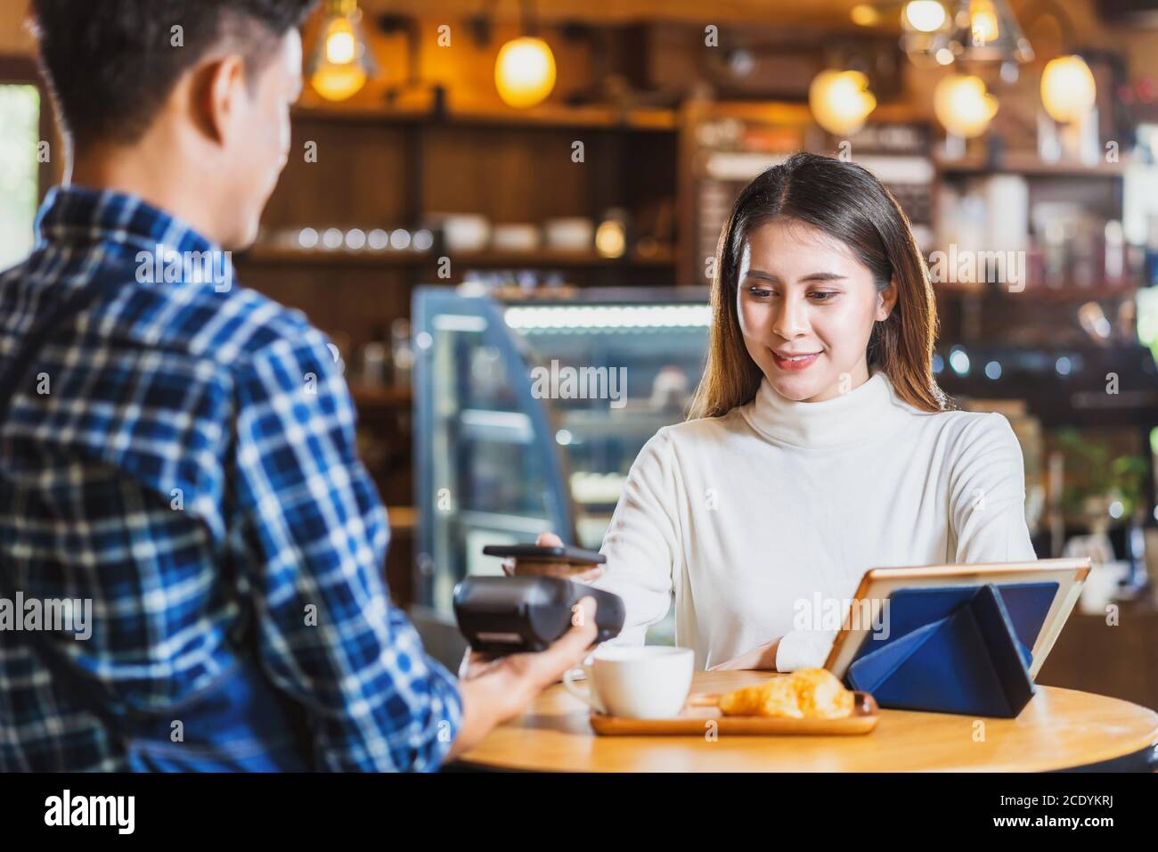 Un cliente asiático que paga con tarjeta de crédito a través de la tecnología nfs sin contacto a un barista asiático del dueño de una pequeña empresa en la mesa de la cafetería, pequeña Foto de stock