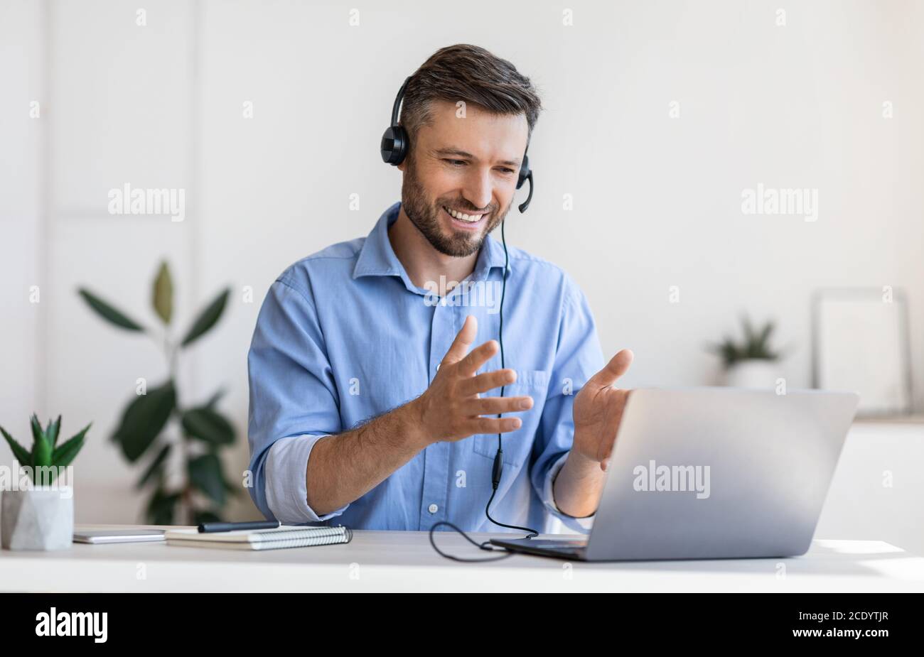 Videoconferencia. Un empleado de la oficina de hombre amable con auriculares que tiene llamada en el ordenador portátil Foto de stock