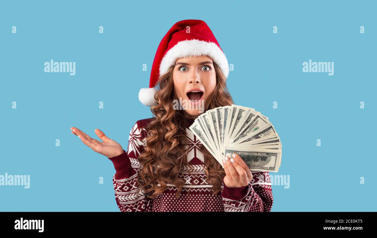 Oferta especial de Navidad. Chica milenaria conmocionada en traje de invierno sosteniendo fan de dinero sobre fondo azul Foto de stock