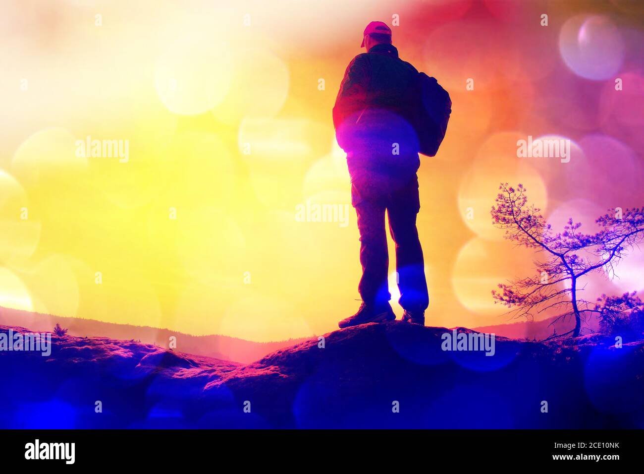 Grano de película. Silueta del hombre con gorra roja y polos en la mano. Hombre con una mochila grande en la roca. Soleado amanecer de primavera en roc Foto de stock