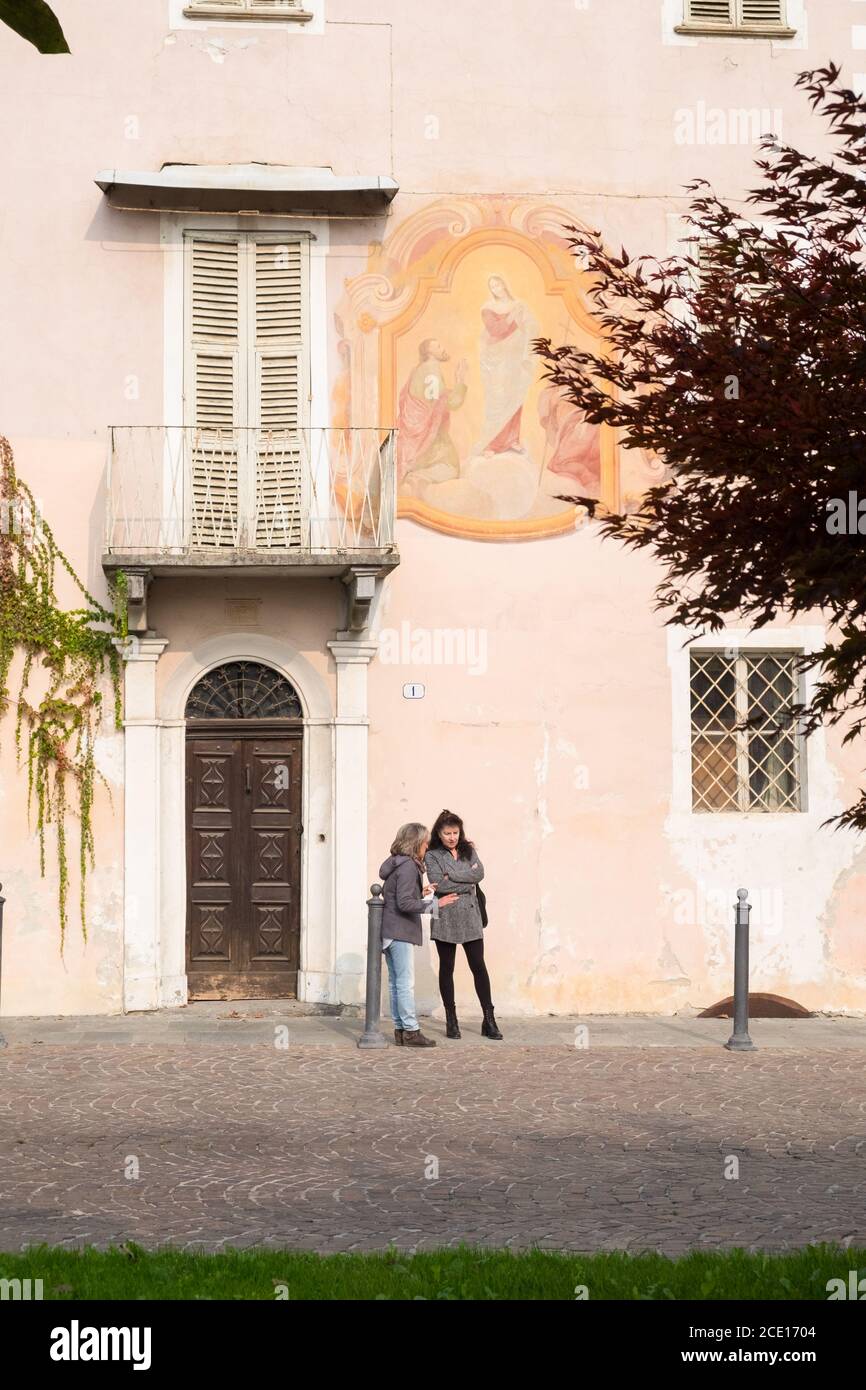 Dos mujeres hablando junto a un edificio tradicional con mural religioso en la pequeña ciudad de Bene Vagienna, Cuneo, Italia Foto de stock