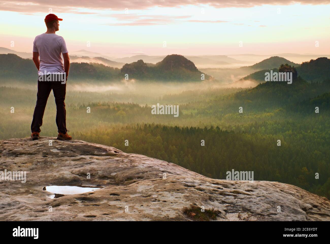 Hombre alto con camisa blanca y pantalones negros con gorra roja de béisbol se mantienen en un acantilado agudo y observan el valle. Colorido morn brumoso Foto de stock