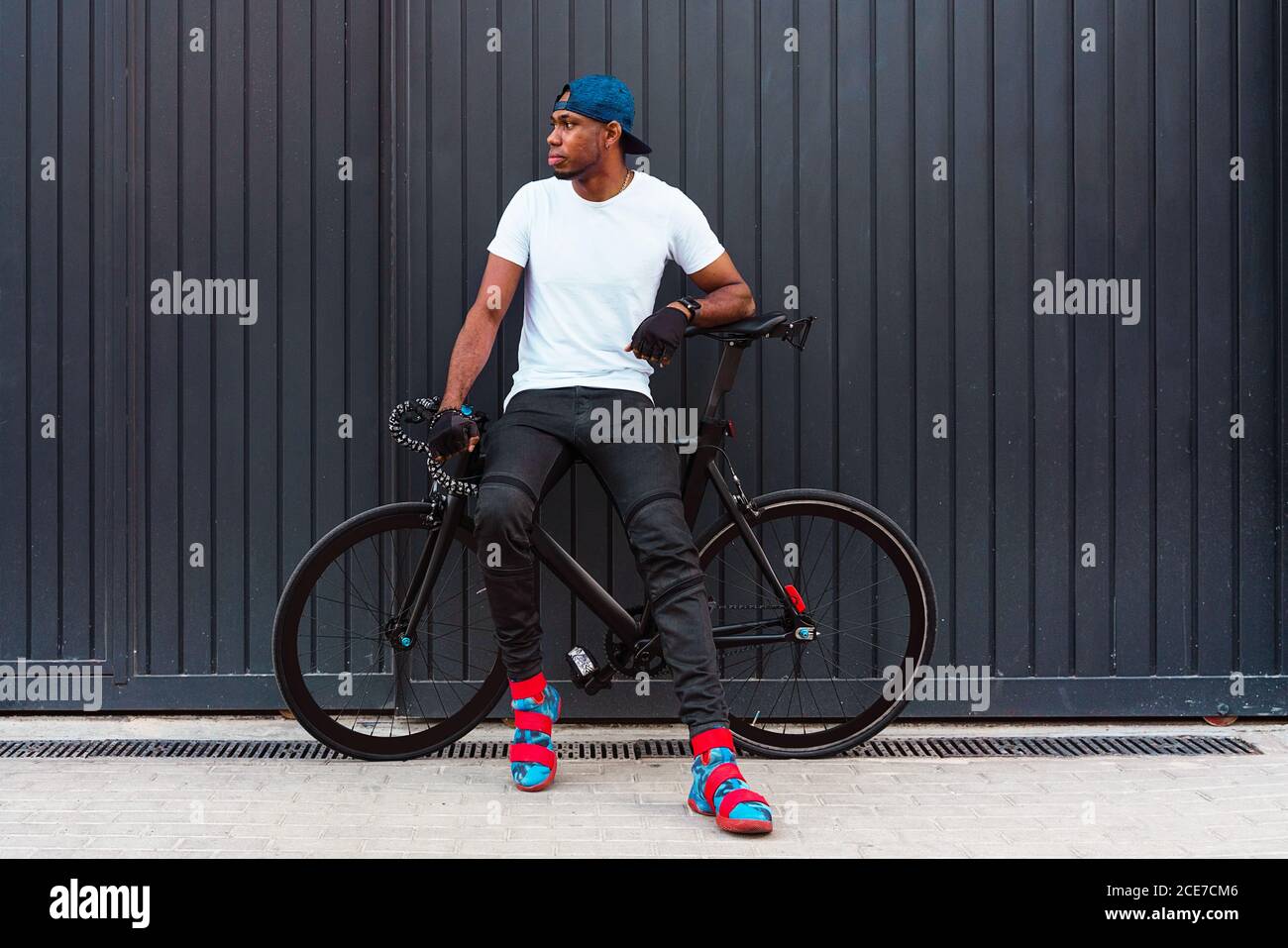 Cuerpo entero de un joven afroamericano serio en casual estilo urbano y  botas de moda de pie con la bicicleta contra pared gris en la calle y  mirando hacia fuera Fotografía de