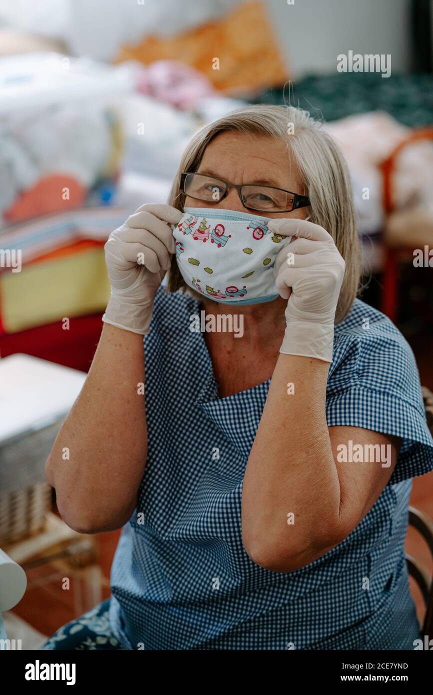 Mujer artesana anciana positiva en anteojos y guantes de látex poner en la  máscara de tela hecha a mano para la prevención del coronavirus mientras  que se sienta a. lugar de trabajo