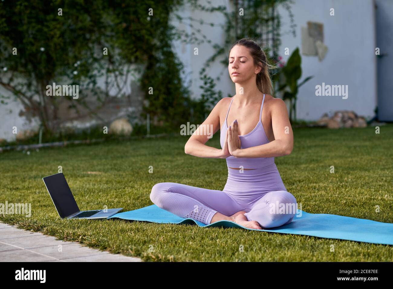 Mujer pacífica de uso activo sentada en Padmasana con Namaste gesto mientras medita y practica yoga en el césped usando la computadora para las clases Foto de stock