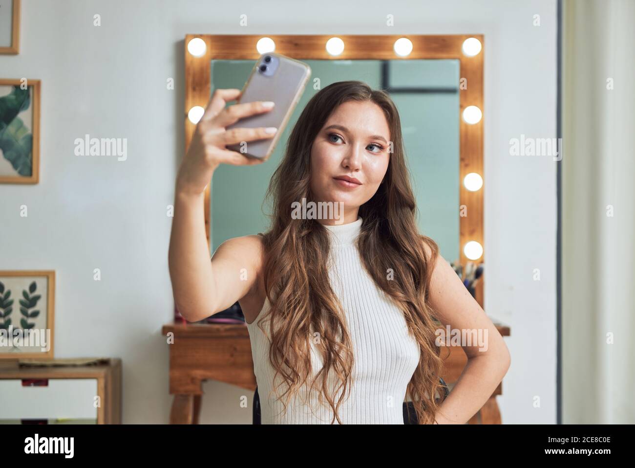 Mujer joven positiva con estilo peinado y maquillaje tomando selfie en el  teléfono móvil moderno contra la mesa de tocador y el espejo con luz  Fotografía de stock - Alamy