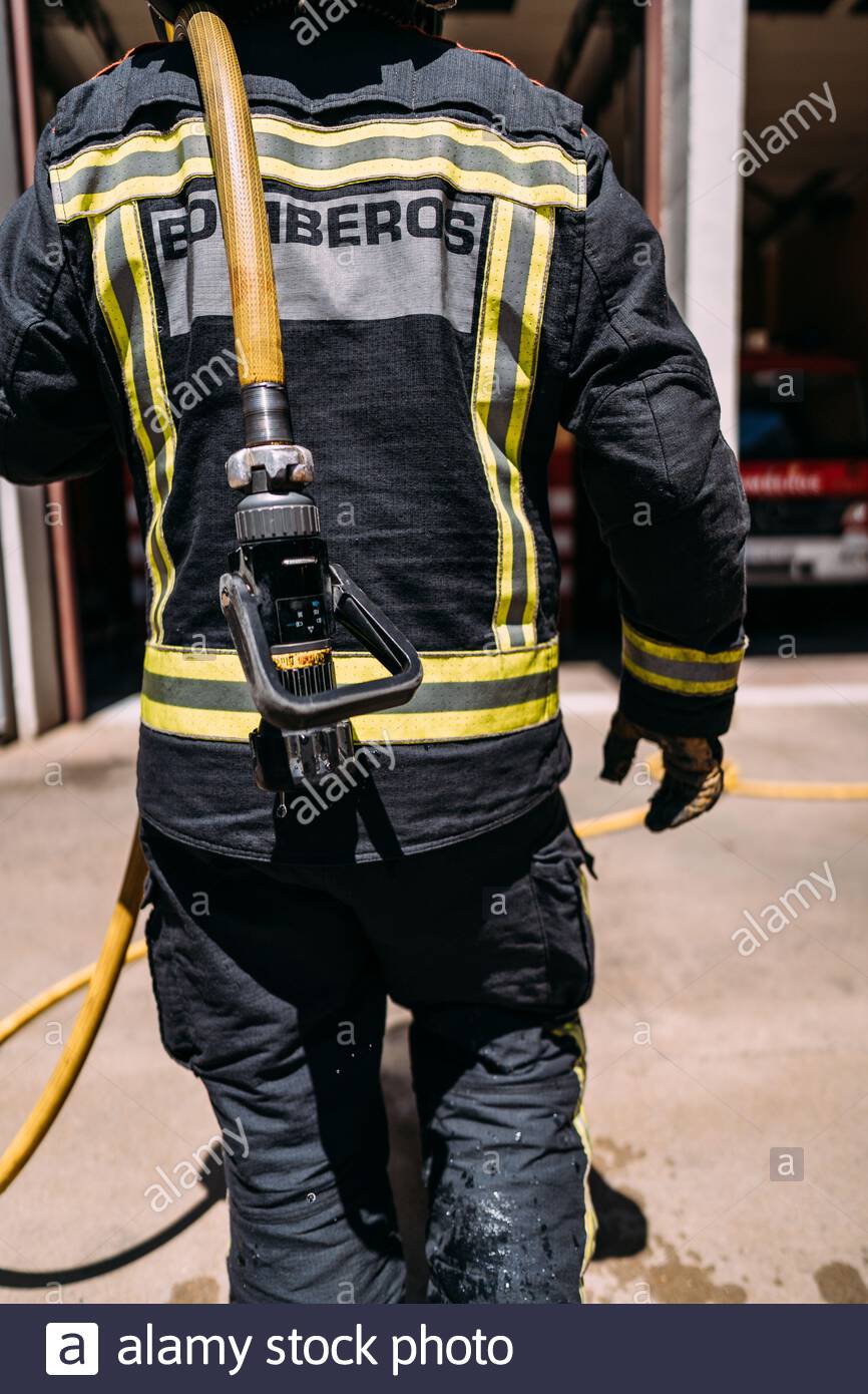 Vista posterior de la cosecha de bomberos anónimos en uniforme de protección con líneas amarillas de pie con manguera larga goteando agua en el pavimento en la luz del día Foto de stock