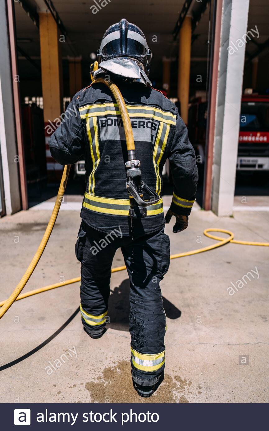 Vista posterior de la cosecha de bomberos anónimos en uniforme de protección con líneas amarillas de pie con manguera larga goteando agua en el pavimento en la luz del día Foto de stock