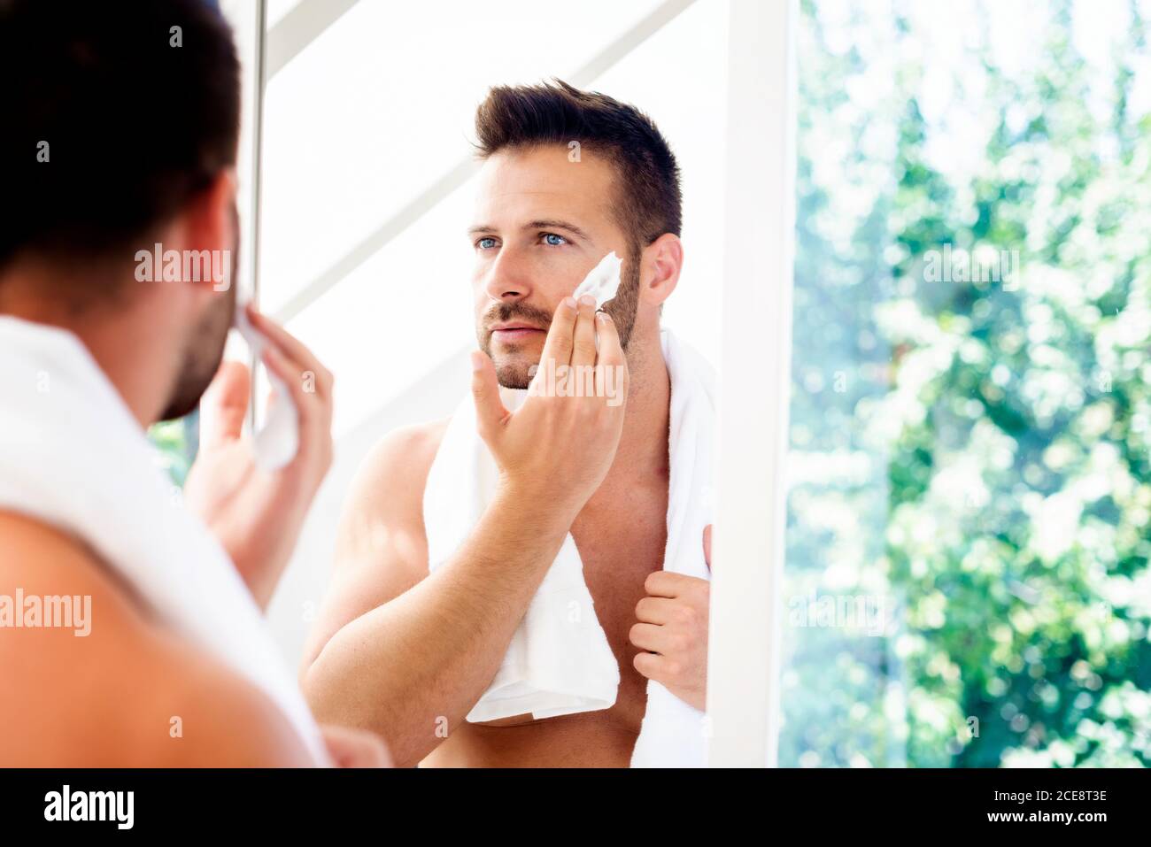 Primer plano de hombre guapo parado en el espejo de la habitación y aplicando espuma de afeitar mientras se prepara el afeitado. Foto de stock