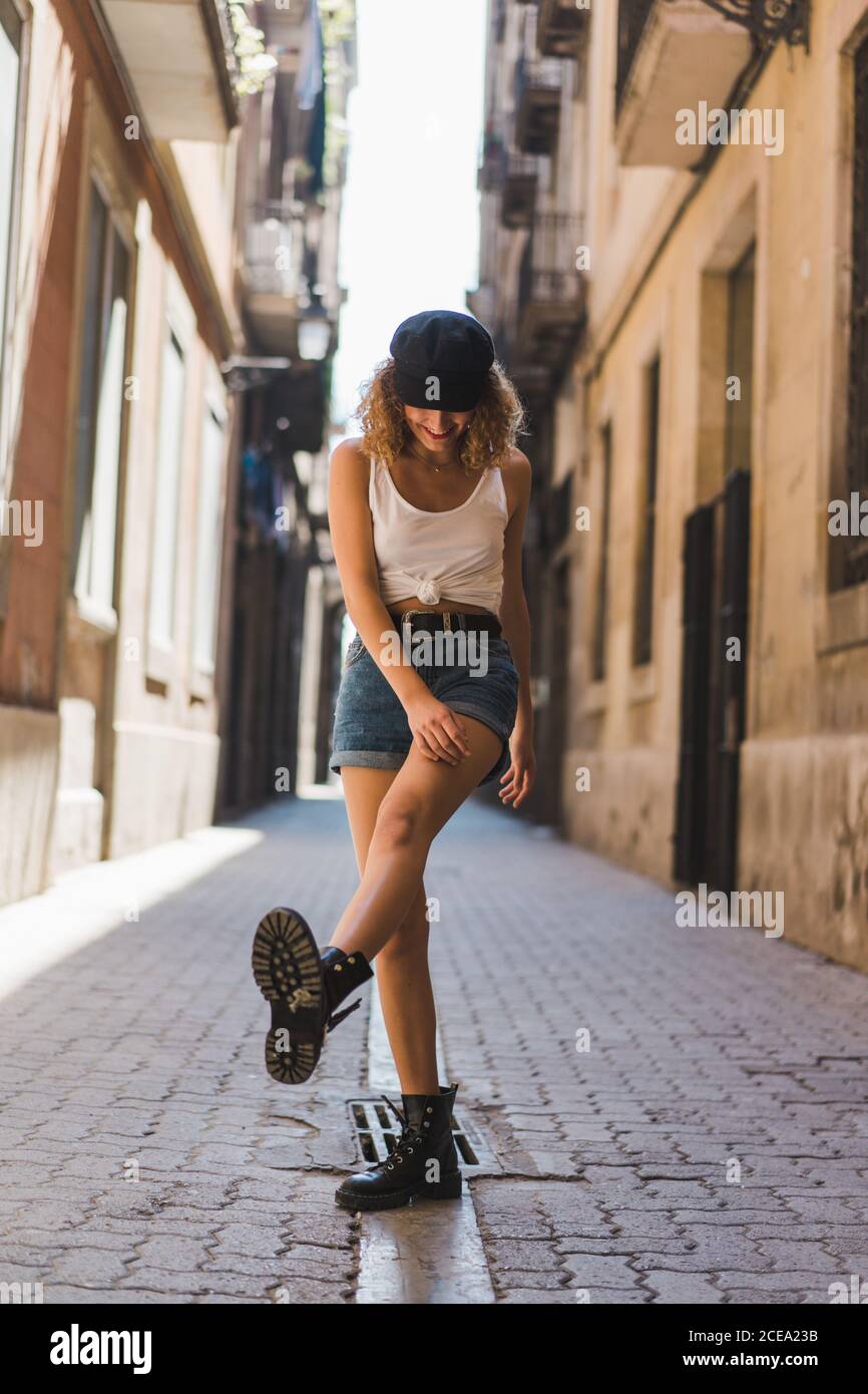 Encantadora mujer rizada con pantalones cortos y botas brutales pie juguetonamente en la calle pavimentada divertirse Fotografía de -