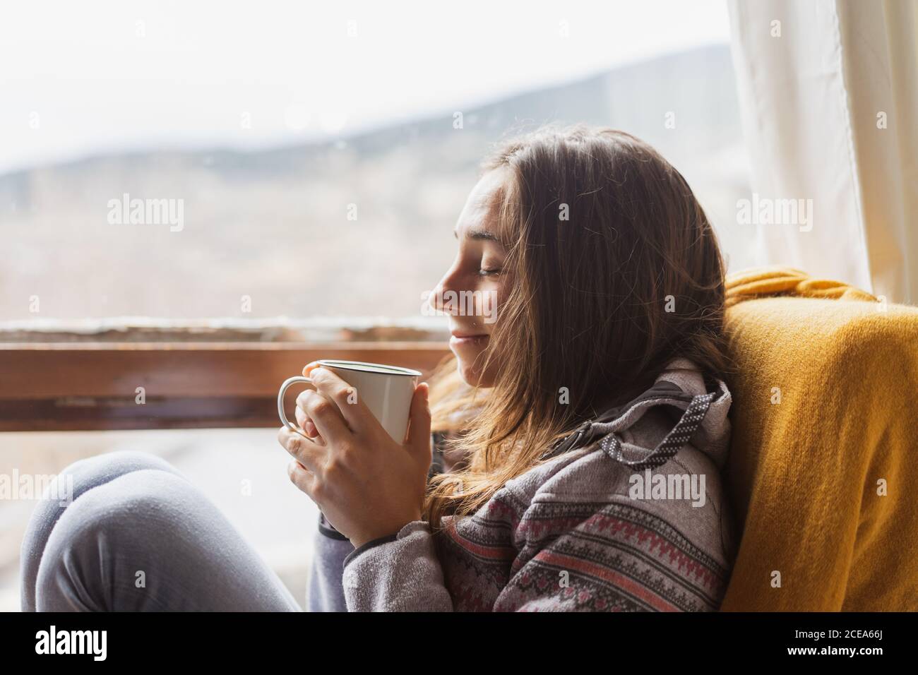 Vista lateral de la linda mujer en un cálido Jersey sonriendo y.. disfrutar de una bebida caliente mientras se sienta en un sillón cerca de enorme ventana en casa Foto de stock
