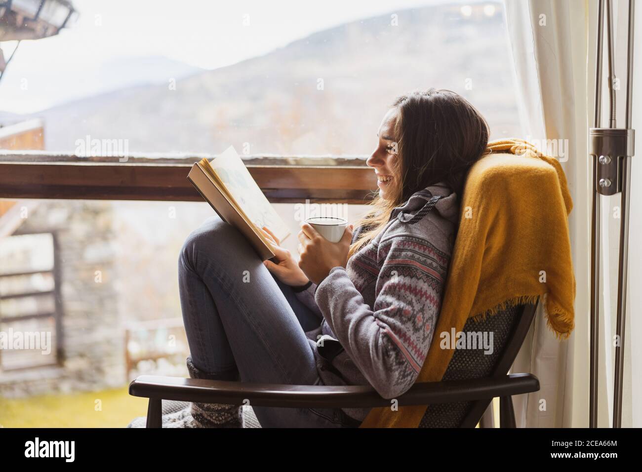 Vista lateral de la señorita con taza de fresco caliente beba leyendo un libro interesante mientras está sentado cerca de una ventana enorme habitación acogedora Foto de stock