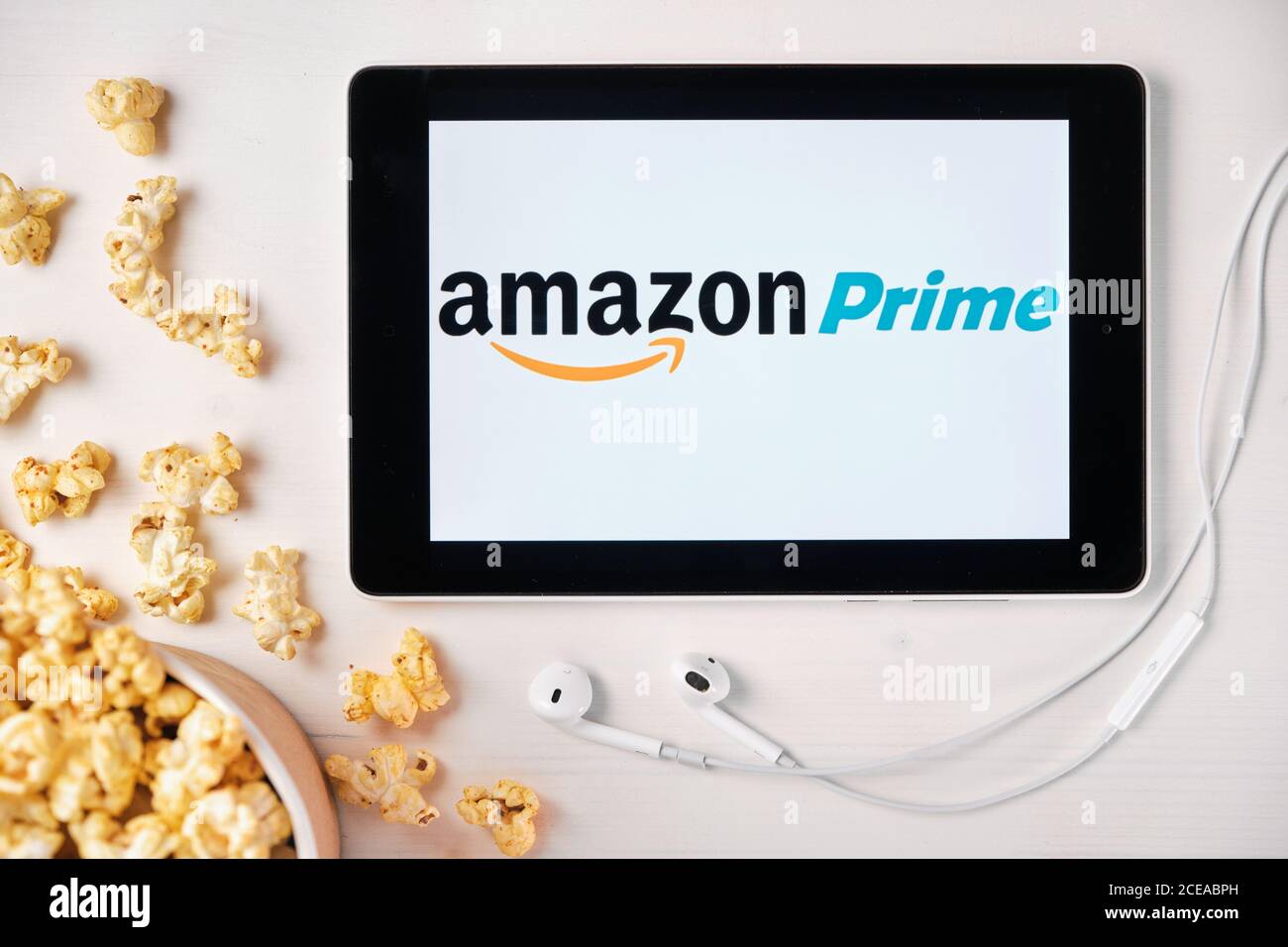 Logotipo de Amazon prime video en la pantalla de la tableta sobre la mesa  blanca y palomitas de maíz rociadas en ella. Auriculares de Apple cerca de  la tableta, agosto Fotografía de