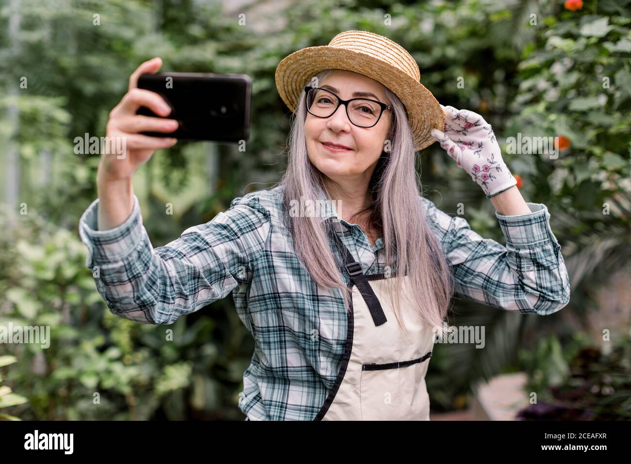 Retrato de atractiva jardinera alegre de 60 años con pelo largo gris, con camisa de cuadros, delantal y sombrero de paja, de pie en un hermoso invernadero Foto de stock