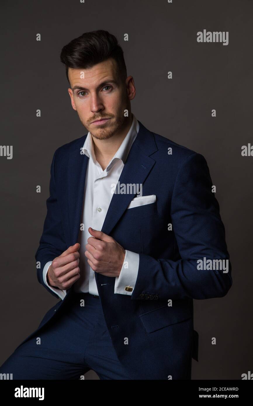 Elegante hombre serio vestido azul oscuro traje con camisa blanca y mirando  la cámara sentado en gris Fotografía de stock - Alamy