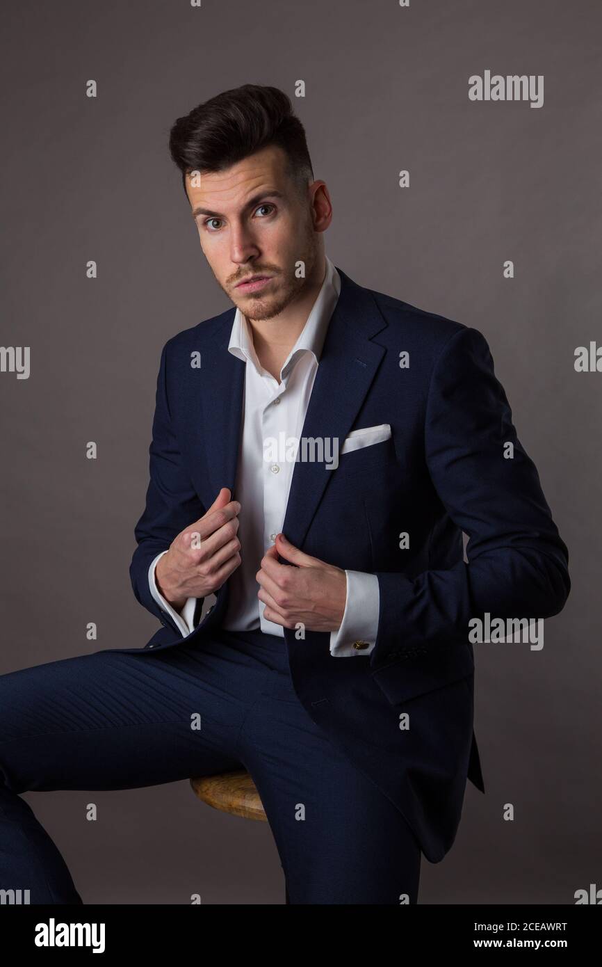 Elegante hombre serio vestido azul oscuro traje con camisa blanca y mirando  la cámara sentado en gris Fotografía de stock - Alamy