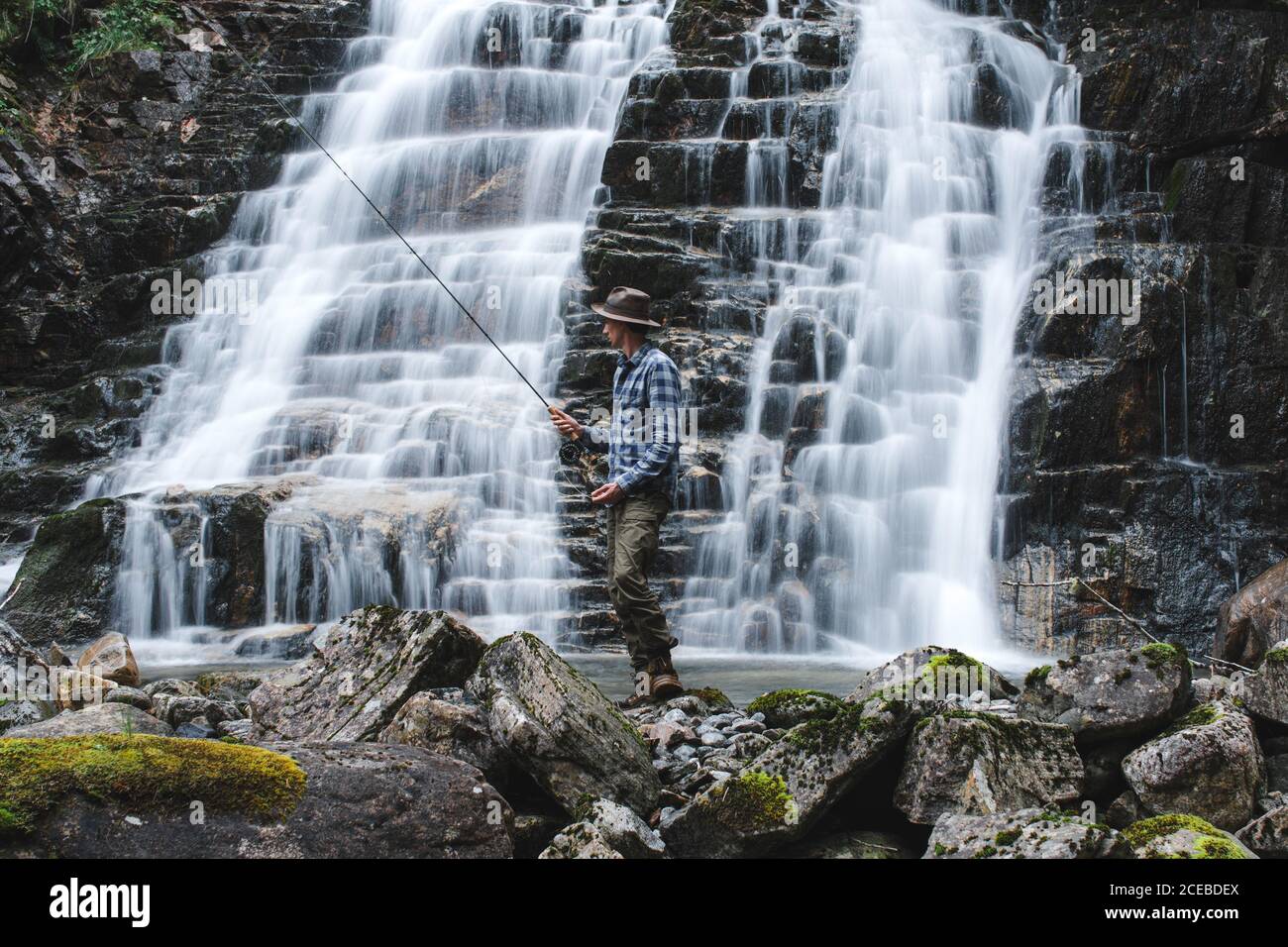 Pescador vestido con sombrero de pesca con camisa azul y gris pantalones de marchar sosteniendo una barra que gira de pie en las rocas detrás la pendiente de una cascada Foto de stock