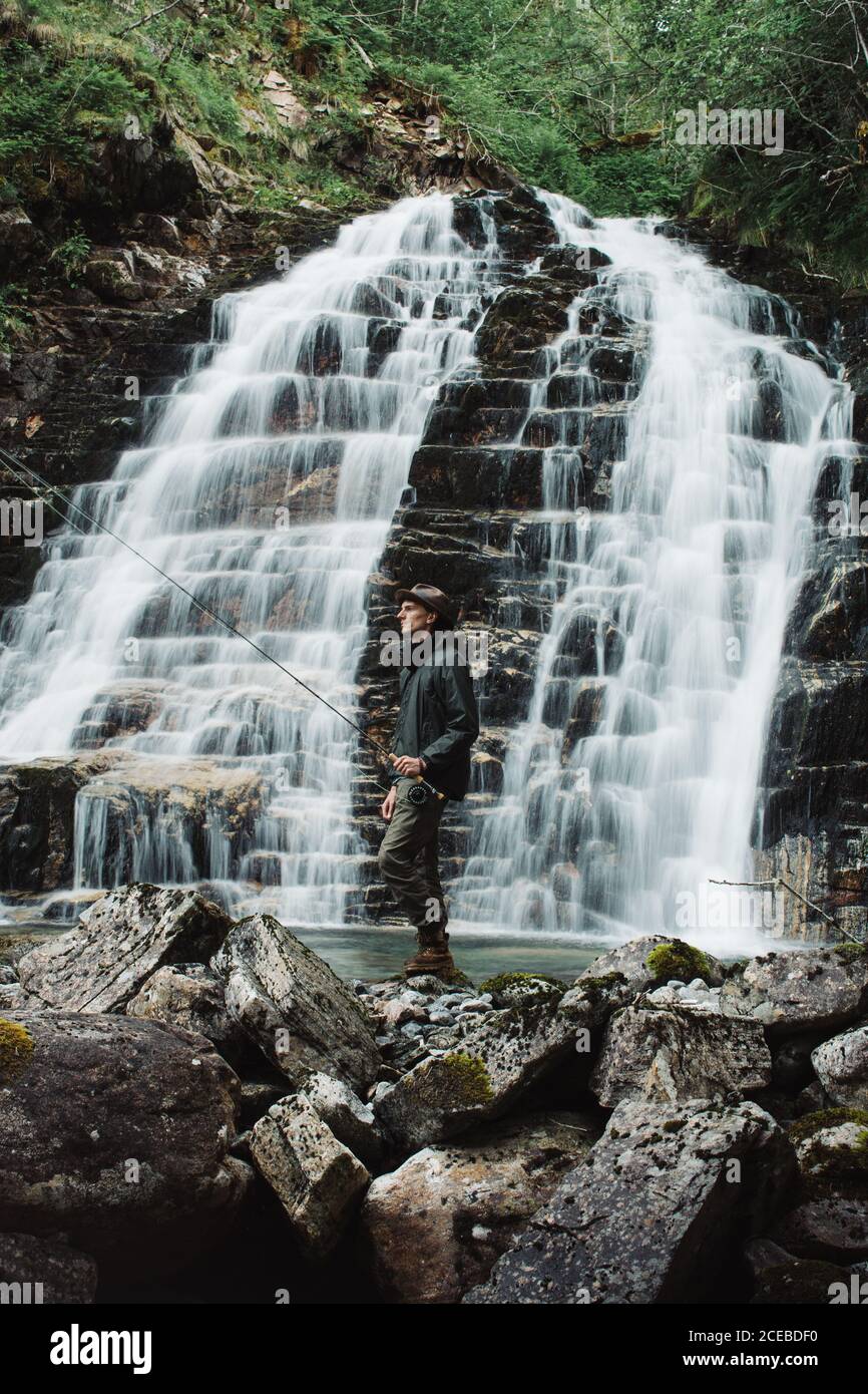 Pescador vestido con sombrero de pesca con camisa azul y gris pantalones de marchar sosteniendo una barra que gira de pie en las rocas detrás la pendiente de una cascada Foto de stock