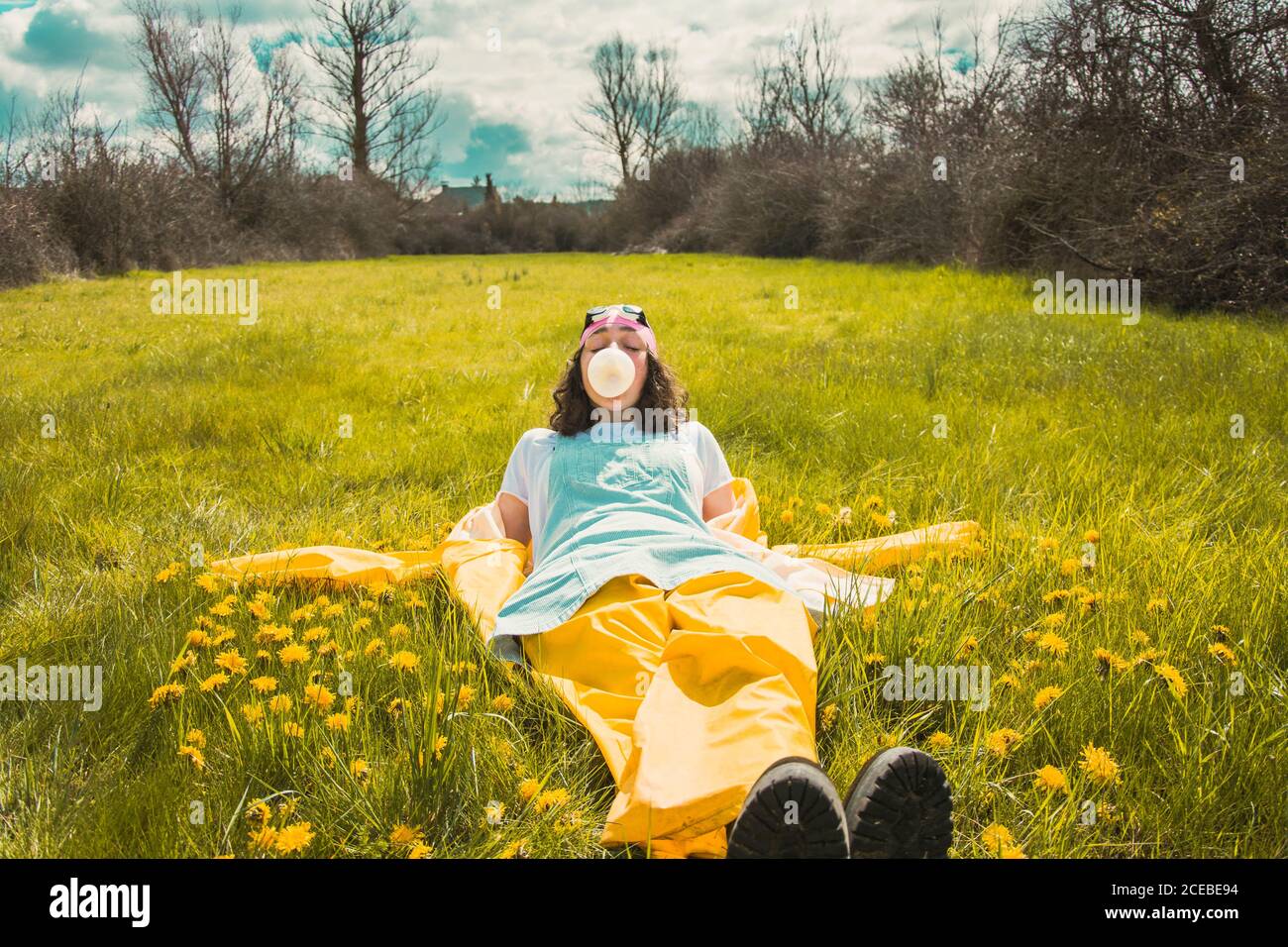 Joven mujer con estilo en chubasquero y sombrero tumbado en prado verde con flores y soñando con la luz del sol Foto de stock