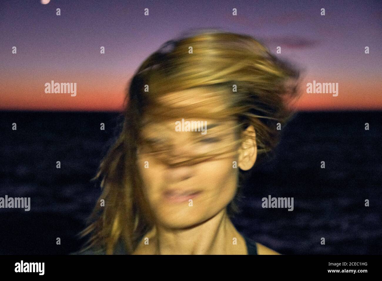 Joven mujer sacudiendo el cabello rubio mientras está de pie contra la noche oscura cielo y mar maravilloso Foto de stock
