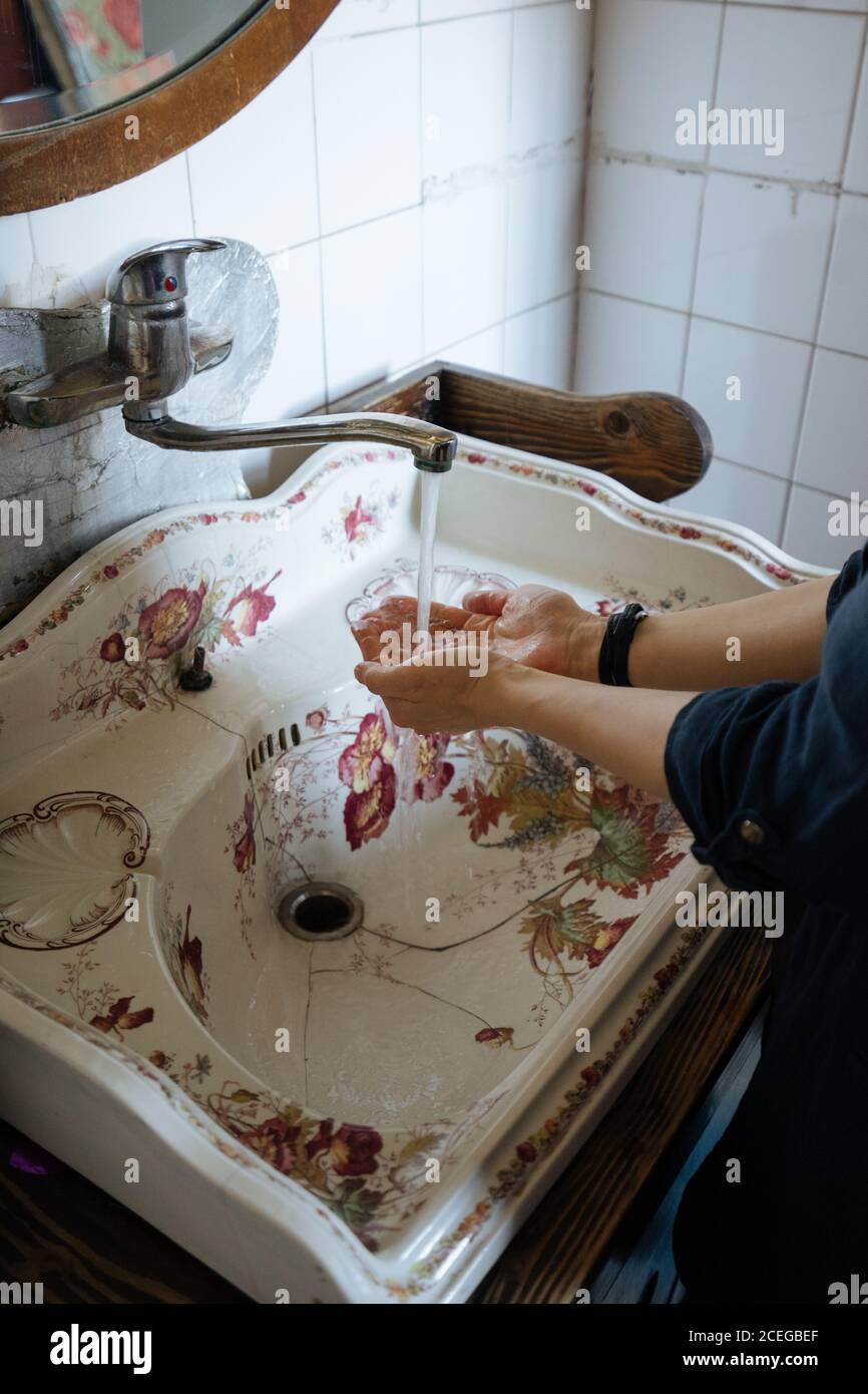 Corta las manos de limpieza femeninas bajo el grifo de agua metálica en el viejo lavabo blanco agrietado con ornamento floral Foto de stock