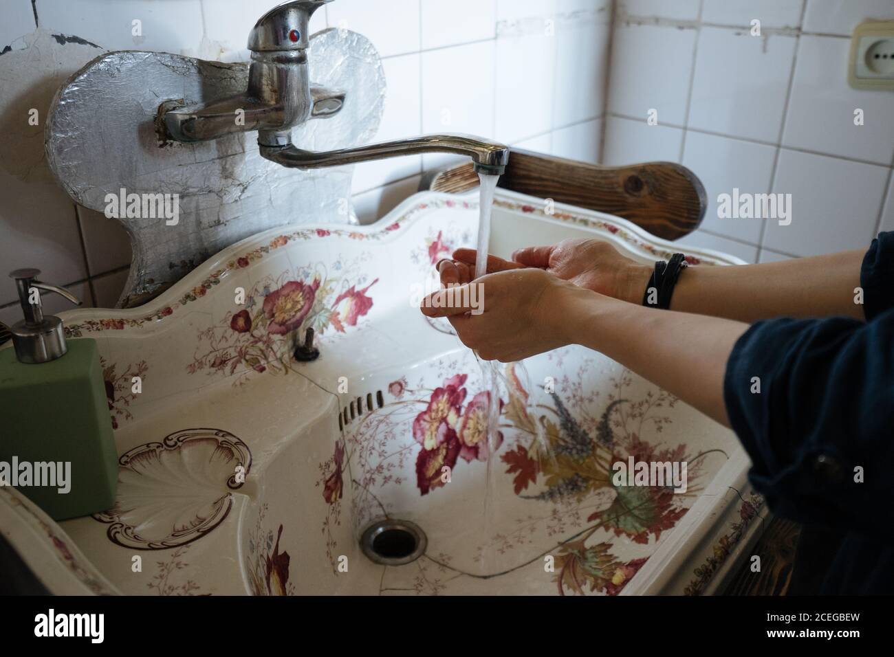 Corta las manos de limpieza femeninas bajo el grifo de agua metálica en el viejo lavabo blanco agrietado con ornamento floral Foto de stock