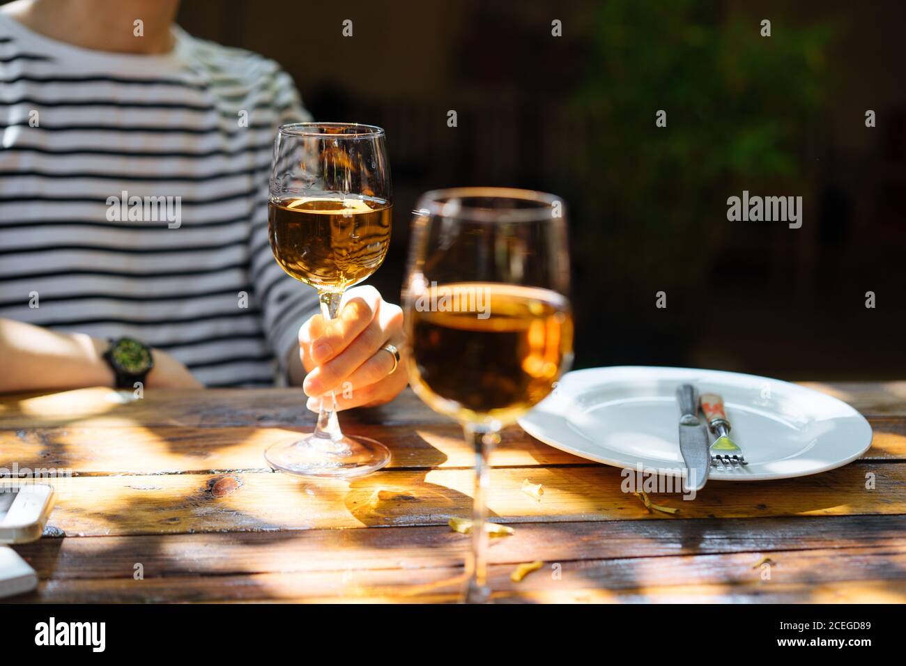 Vista de la hembra en blusa de rayas con elegante cristal de vino blanco  sentado en una mesa de tablas de madera con bebida escucha sobre fondo  borroso Fotografía de stock -