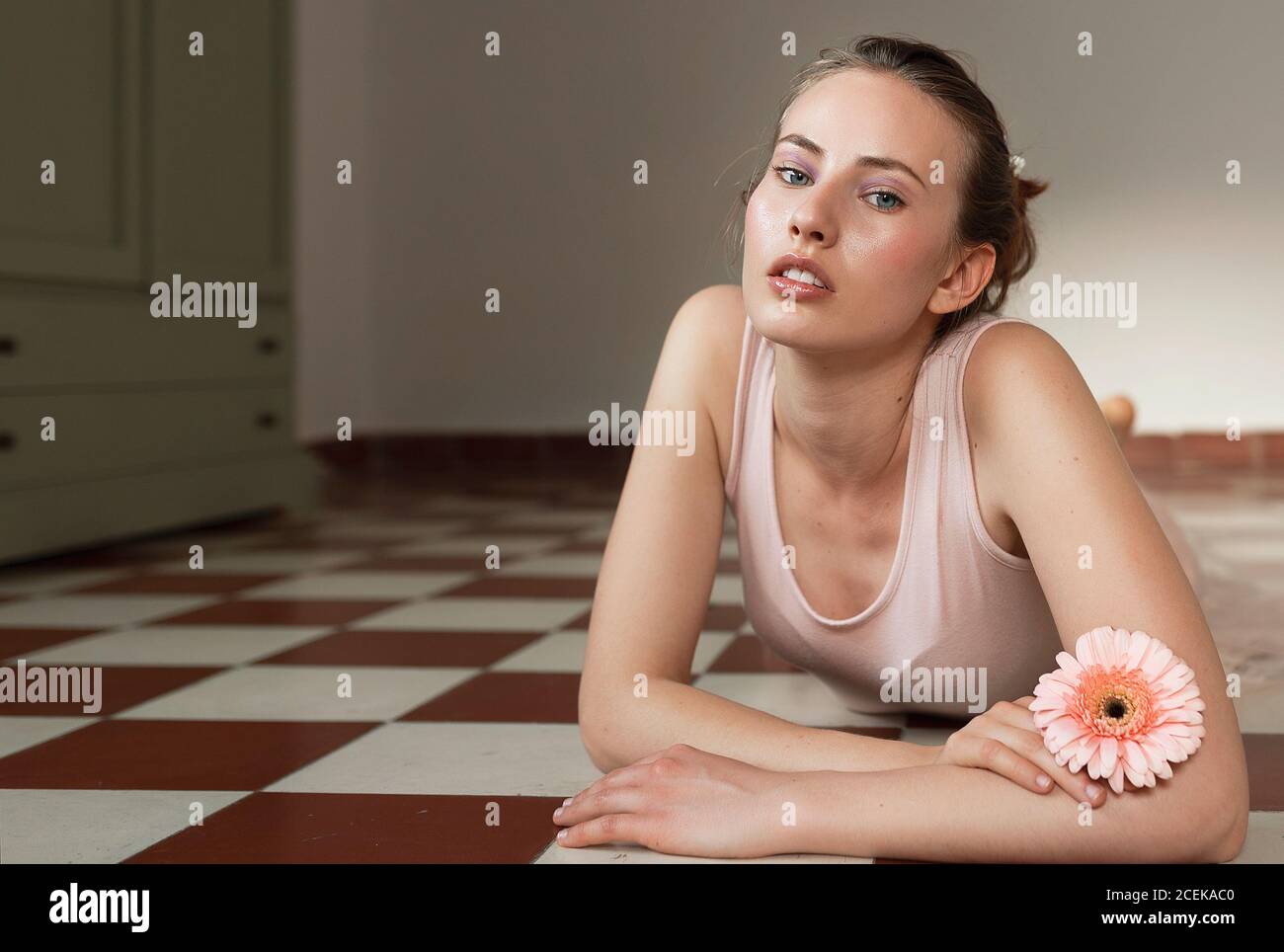 Hermosa joven mujer cruzando las manos, sosteniendo la flor, colocándose en el suelo de baldosa de la cocina y mirando la cámara Foto de stock