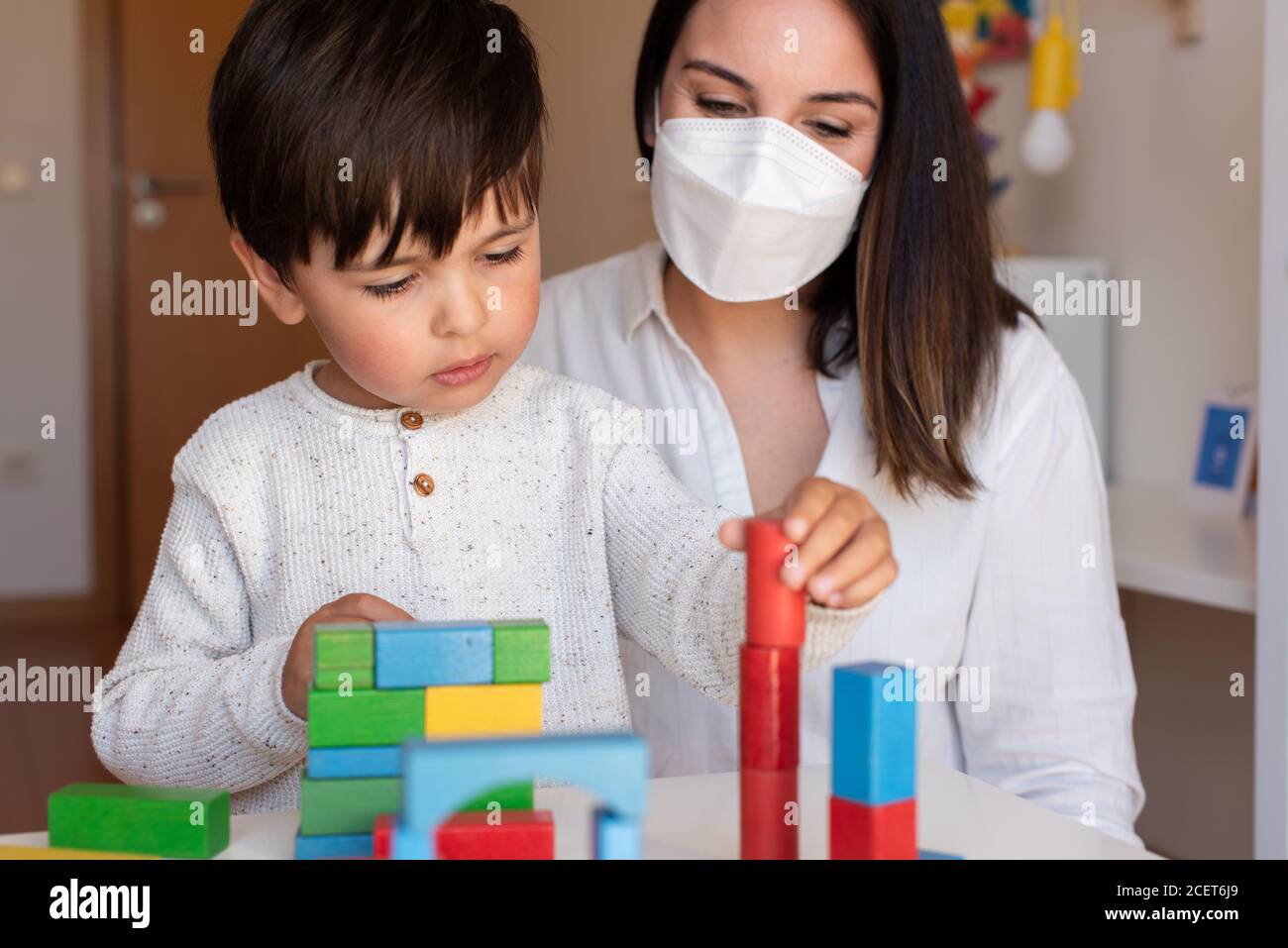Liltle Preschooler Kid jugando con bloques de madera y maestro educador ayuda a usar la máscara para la pandemia del coronavirus. Resolución de problemas en casa. Comunidad de aprendizaje. Foto de stock