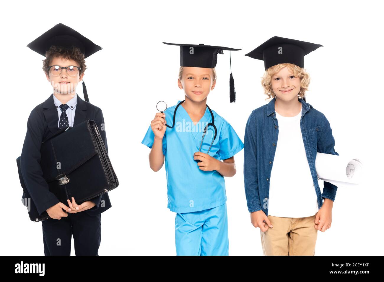 niños en gorras de graduación vestidos con trajes de diferentes profesiones con planos, estetoscopio y maletín aislados en blanco Foto de stock