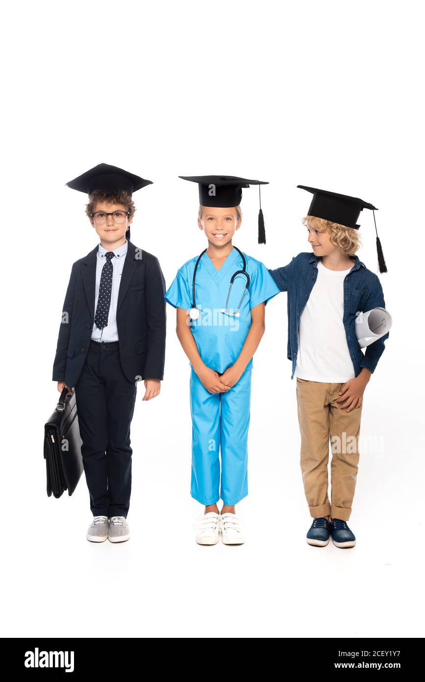 niños en gorras de graduación vestidos con trajes de diferentes profesiones de pie con blueprint, maletín y estetoscopio aislados en blanco Foto de stock