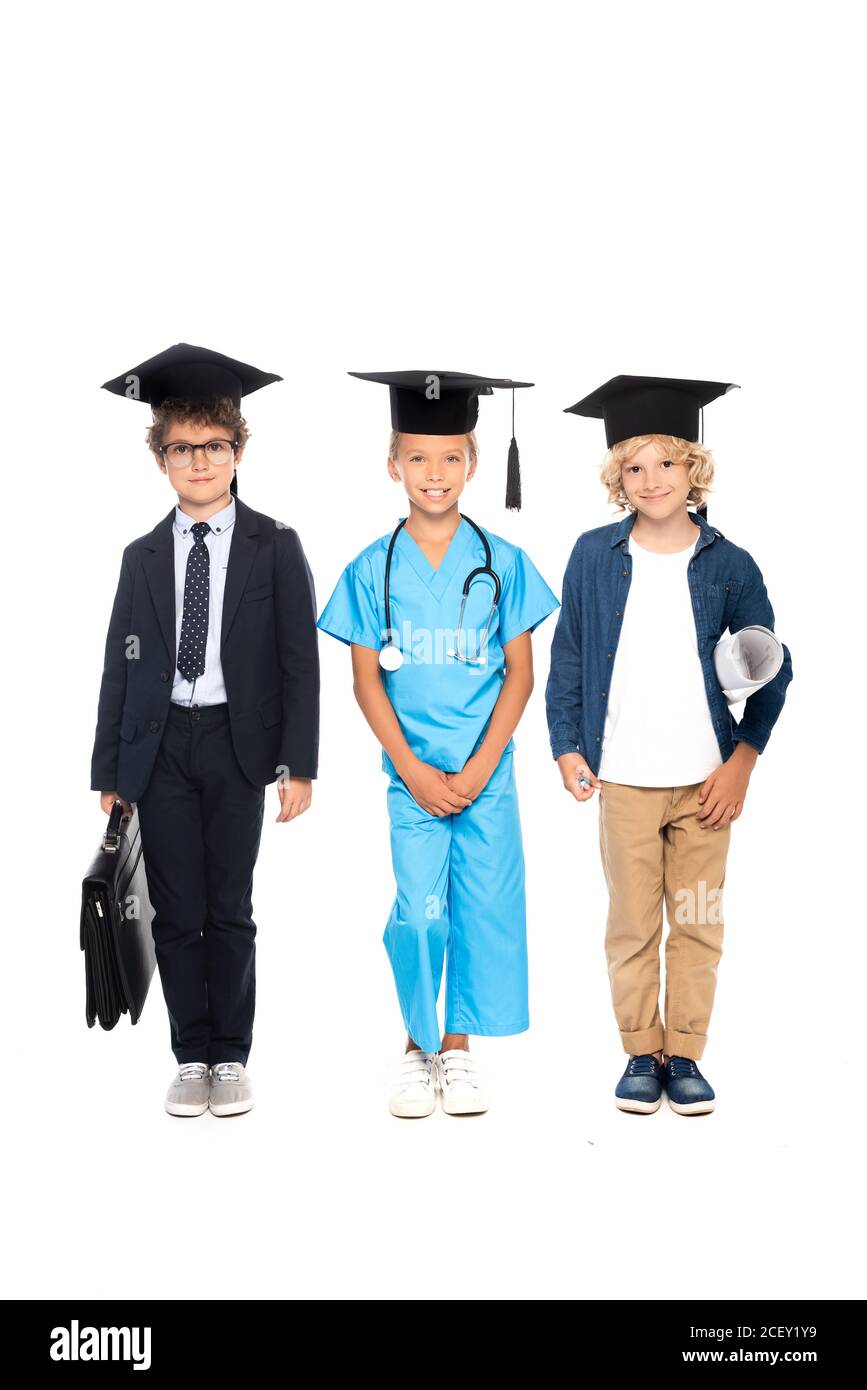 niños en gorras de graduación vestidos con trajes de diferentes profesiones de pie con estetoscopio, blueprint y maletín aislados en blanco Foto de stock