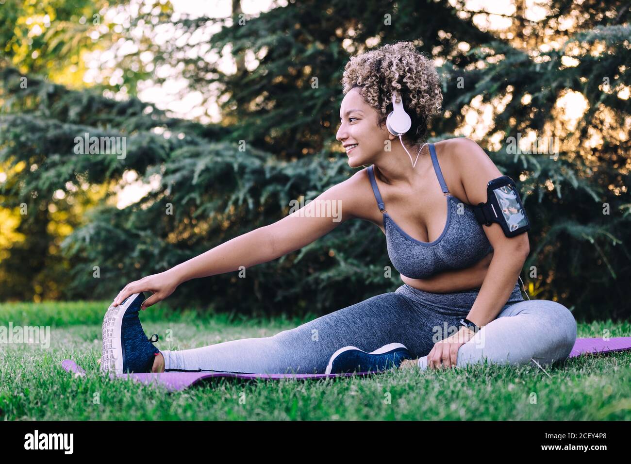 Mujer étnica joven feliz con pelo rizado en sportswear escuchar a la música con auriculares y smartphone y haciendo sentado hacia delante doble el ejercicio mientras estira el cuerpo durante el entrenamiento de fitness en verano parque Foto de stock