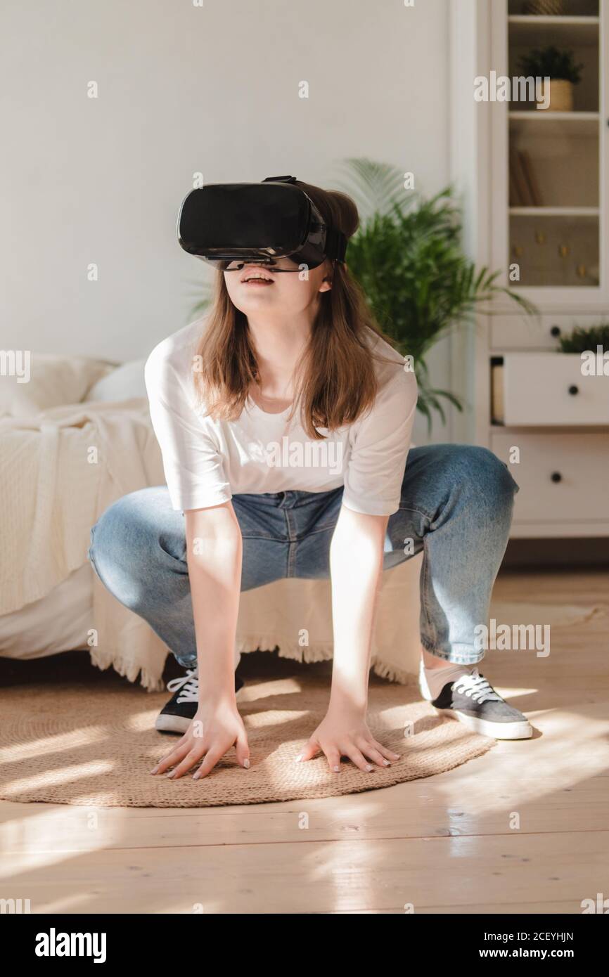 Mujer joven en jeans y camiseta blanca con casco de realidad virtual juega,  trabaja, mira video en la habitación al lado de la cama. Concepto de  tecnolo moderno Fotografía de stock -