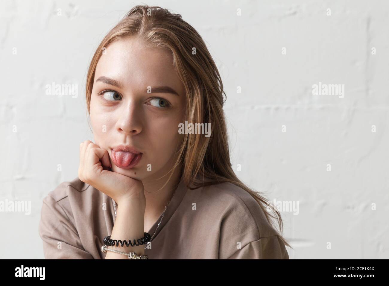 Chica adolescente rubia divertida muestra lengua, primer plano retrato de estudio con luz natural sobre pared blanca Foto de stock