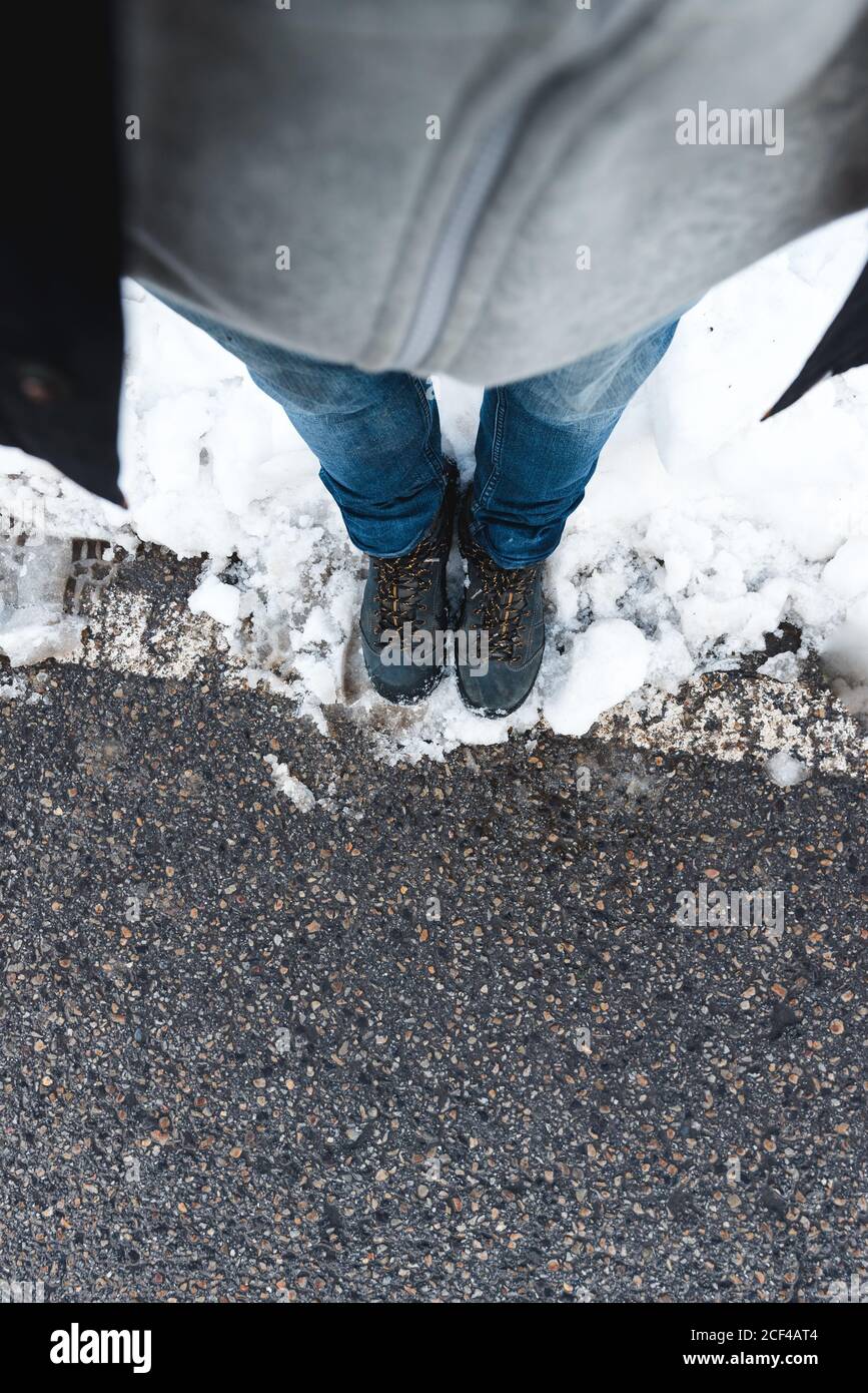 Vista superior de la cosecha irreconocible persona en jeans y trekking zapatos de pie en asfalto carretera nevada durante el día Foto de stock