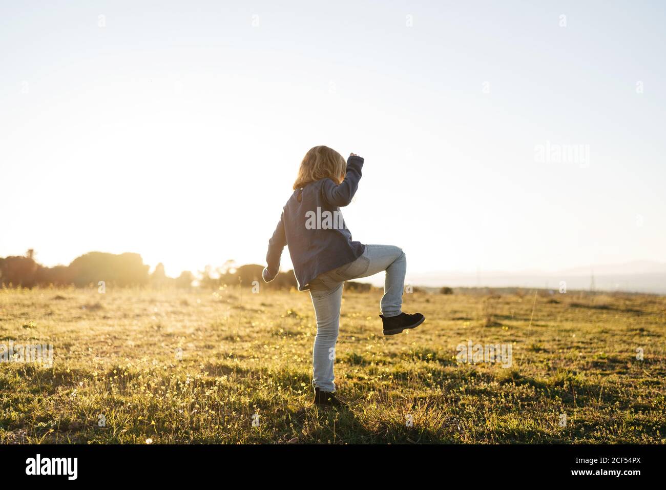 Vista posterior de la adorable chica activa anónima en informal usa jugando y bailando en el campo verde mientras disfrutas de sol noche de verano en el campo Foto de stock