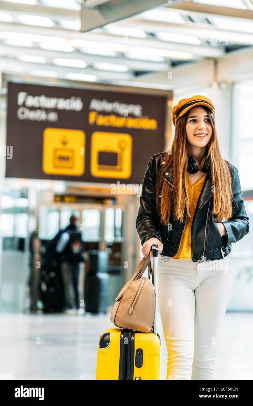 Mujer sonriente con estilo en gorra amarilla chaqueta de cuero negro y  blusa amarilla con auriculares de pie con maleta en el aeropuerto moderno  terminal y mirando la cámara Fotografía de stock -