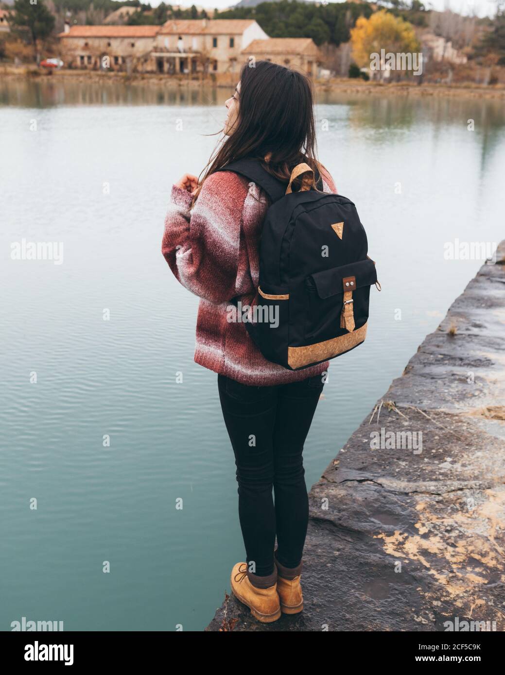 Vista posterior de la joven mujer viajera activa en conjunto informal con  la mochila de pie cerca del tranquilo lago en la ciudad en nuboso día de  otoño Fotografía de stock -