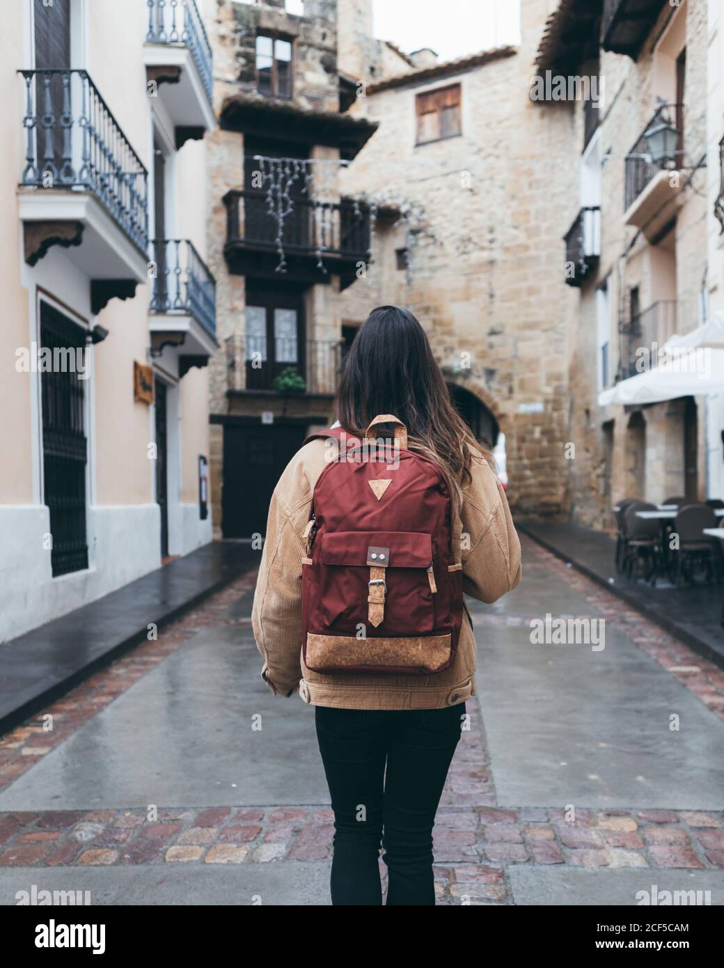 Vista posterior de la joven turista femenina en ropa de abrigo con mochila  a la moda caminando en una calle estrecha con un antiguo edificio de piedra  en la ciudad Fotografía de