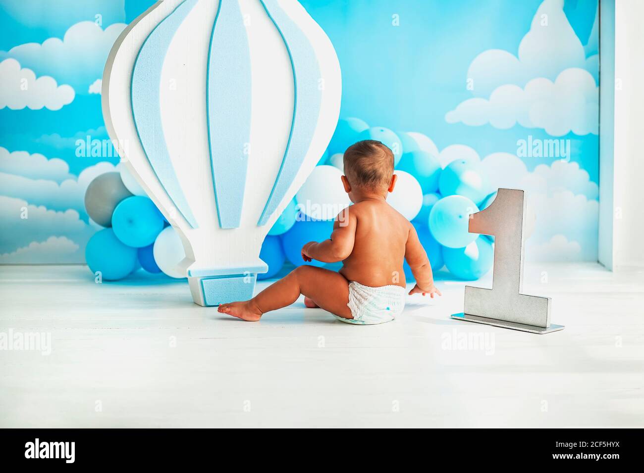 El primer cumpleaños del bebé. Decoración de fiesta con un gran globo y  pequeños globos blancos y azules contra una pared azul con nubes blancas. Un  niño pequeño Fotografía de stock -
