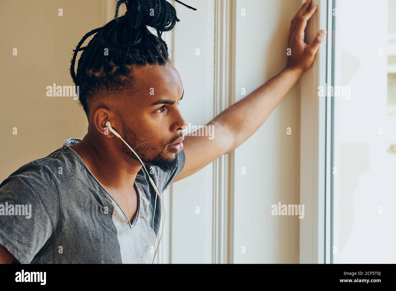 Vista lateral del hombre étnico serio con peinado creativo escuchar a la música y apoyándose en el marco de la ventana Foto de stock