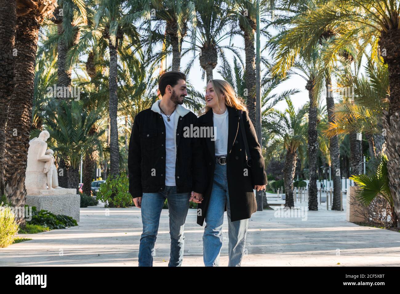 Alegre joven pareja romántica en jeans y chaquetas de moda negro caminar  por el callejón en el parque con palmeras tropicales y disfrutar vacaciones  juntos en un día soleado Fotografía de stock -