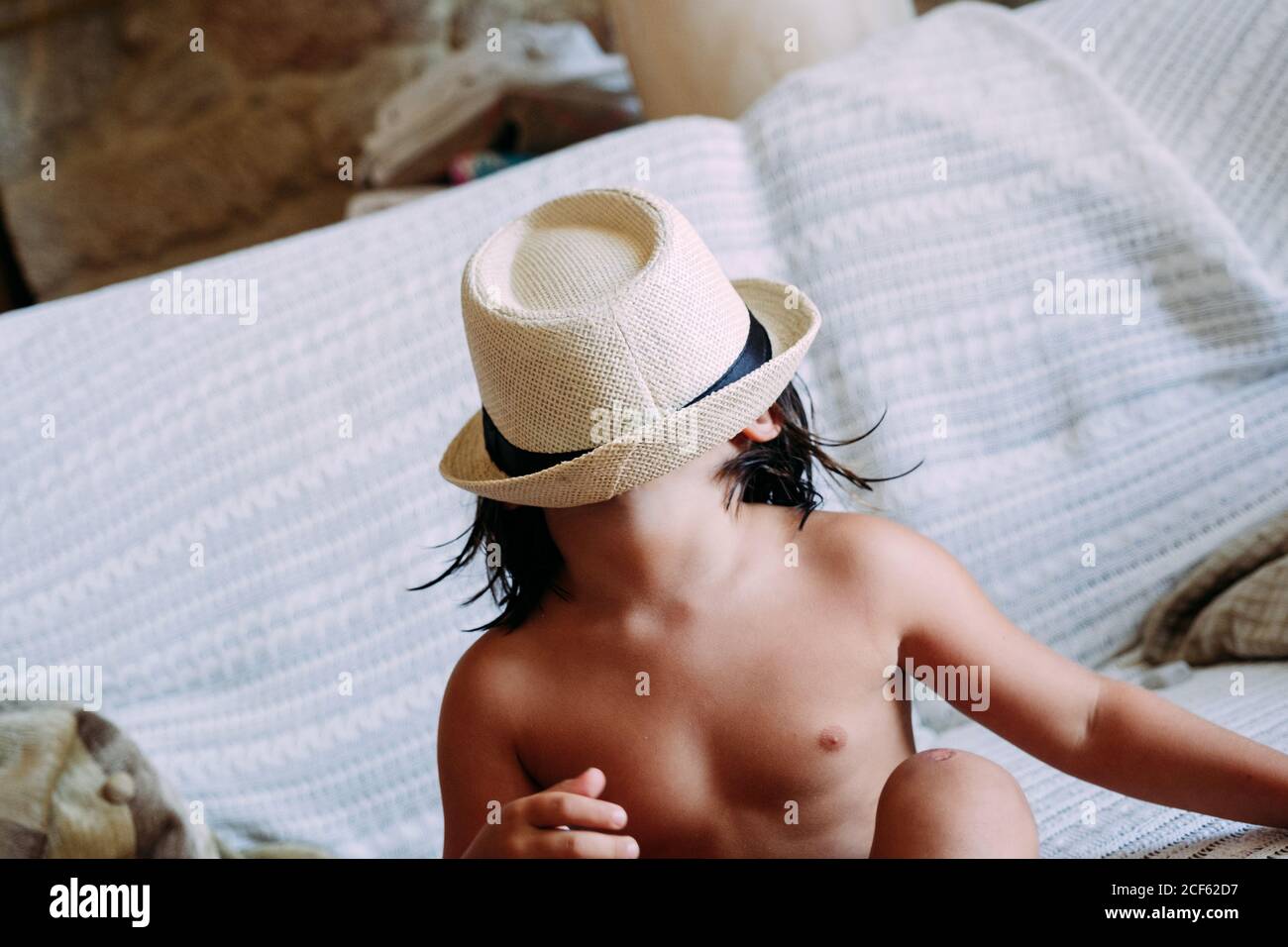 niño anónimo sin ropa que cubra la cara con un sombrero sentado en el sofá  Fotografía de stock - Alamy
