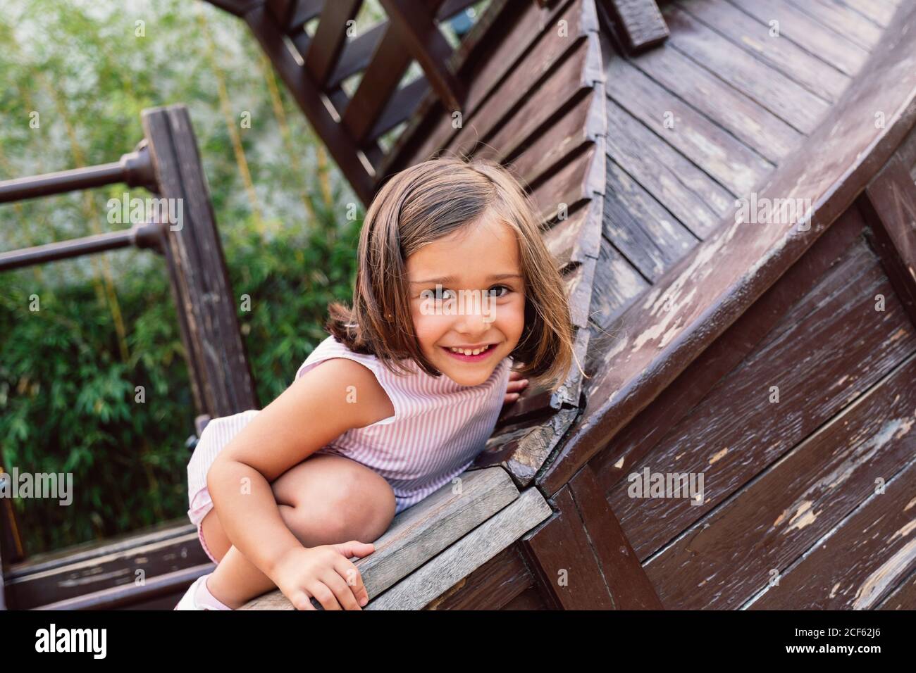 Alegre chica nimble subir a la estructura de madera y mirar cámara con sonrisa Foto de stock