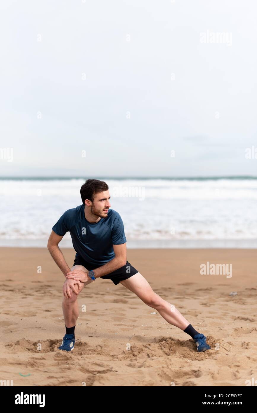 Gimnasta masculina en ropa deportiva estirando las piernas y mirando lejos  en la playa de arena vacía con el mar azul y el cielo encendido fondo  borroso Fotografía de stock - Alamy