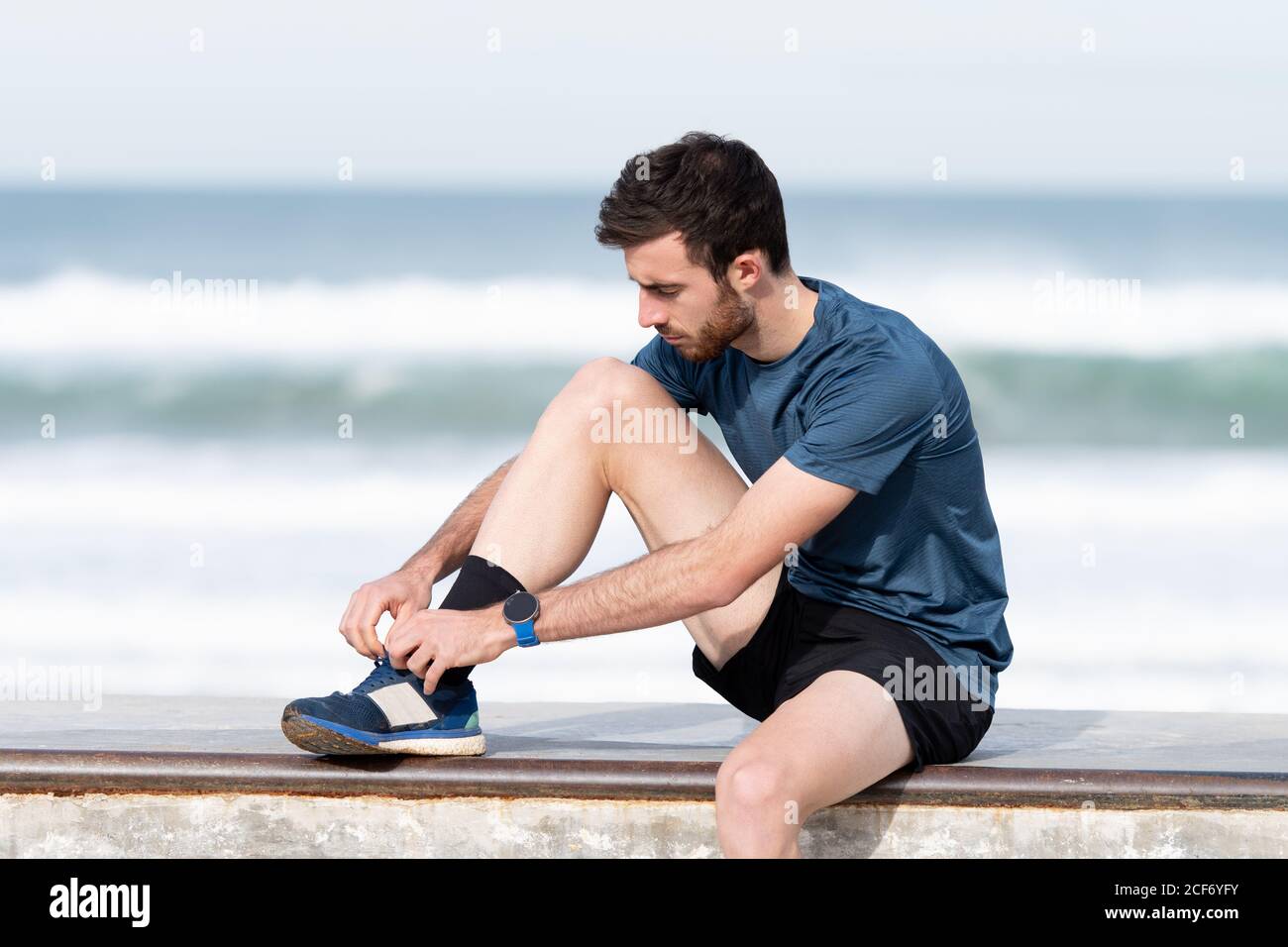 Atleta de pelo negro barba en camiseta azul y.. pantalones cortos en la valla amarrando cordones con el mar azul y el cielo fondo borroso Fotografía de stock -
