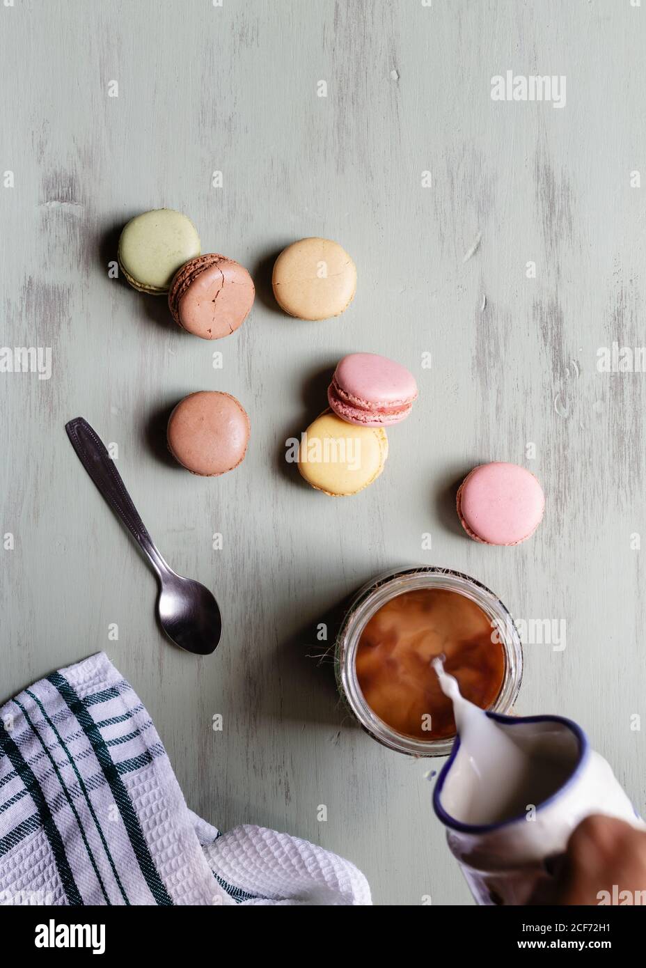De arriba persona anónima vertiendo leche en taza de café cerca de un montón de coloridos macarrones y cuchara con servilleta mesa blanca Foto de stock